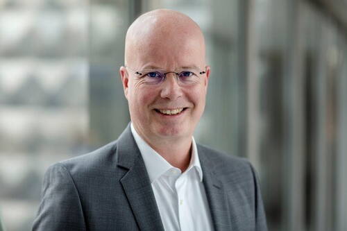 Glatzköpfiger Mann mit Brille im grauen Anzug lächelt freundlich in moderner Büroumgebung