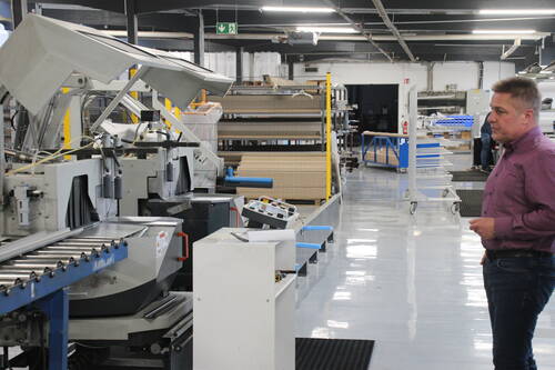 Mann in Arbeitskleidung überwacht eine automatisierte Metallverarbeitungsmaschine in einer aufgeräumten Fabrikhalle mit Regalen und Förderanlagen im Hintergrund.