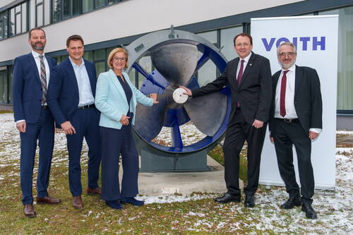 Fünf Geschäftsleute in Anzug und Kostüm posieren im Freien vor einem großen Industriepropeller neben einem VOITH-Firmenbanner.