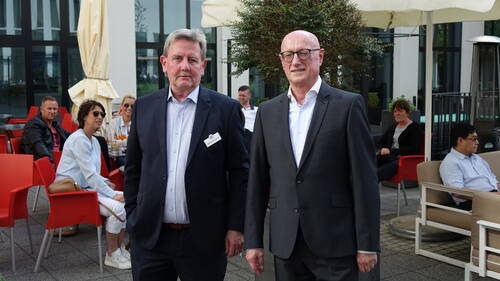 Zwei ältere Männer in Anzügen stehen im Mittelpunkt einer Sitzterrasse, während andere Gäste entspannt auf roten Stühlen im Hintergrund plaudern und Getränke genießen.