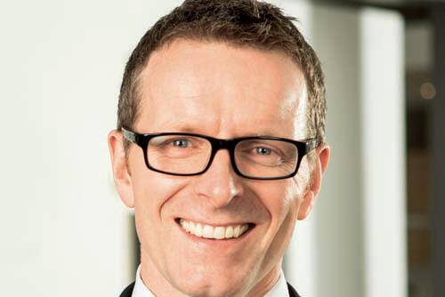 Lächelnder Mann mit kurzer brauner Frisur und schwarzer Brille in Businesskleidung vor unscharfem Bürohintergrund