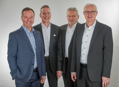 Vier lächelnde Geschäftsmänner mittleren bis höheren Alters posieren im Anzug nebeneinander vor neutralem, hellem Hintergrund.