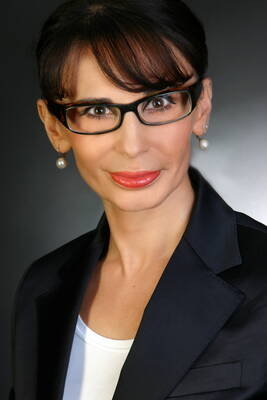 Elegant gekleidete Frau mit dunklem Haar, Brille und Perlenohrringen lächelt selbstbewusst vor dunklem Hintergrund in die Kamera.