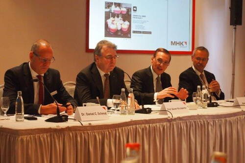 Vier Anzugträger sitzen bei einer Pressekonferenz hinter einem mit Mikrofonen und Namensschildern bestückten Tisch und diskutieren vor einer Präsentationsfolie mit MHK-Logo.