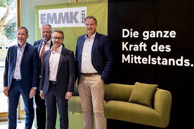 Vier Geschäftsleute in Anzügen posieren vor einem EMMK‑2021‑Banner neben einem schwarzen Hintergrund mit der Aufschrift „Die ganze Kraft des Mittelstands“ und einem grünen Sofa.