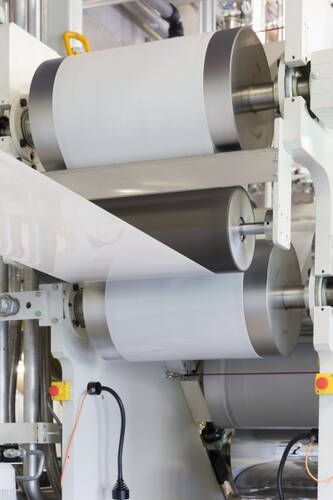 Große industrielle Walzmaschine führt eine breite, glänzende Kunststoff- oder Papierbahn präzise über mehrere Metallrollen