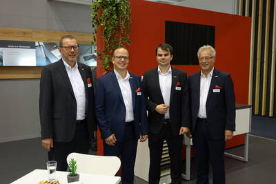 Vier Geschäftsleute in Anzügen mit roten Einstecktüchern posieren lächelnd vor einem modernen, rot-grauen Messestand mit Pflanzen und Präsentationswänden im Hintergrund.