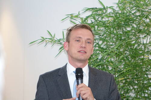 Mann im Anzug spricht in ein Mikrofon vor einem grünen Pflanzenhintergrund