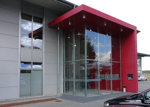 Moderner Eingangsbereich eines Gewerbegebäudes mit großer Glasfassade und markantem roten Vordach auf grauer Metallverkleidung