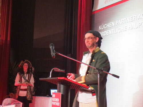 Moderator im Piratenkostüm spricht am Rednerpult auf einer Bühne, während eine ebenfalls als Piratin verkleidete Frau lächelnd im Hintergrund steht.
