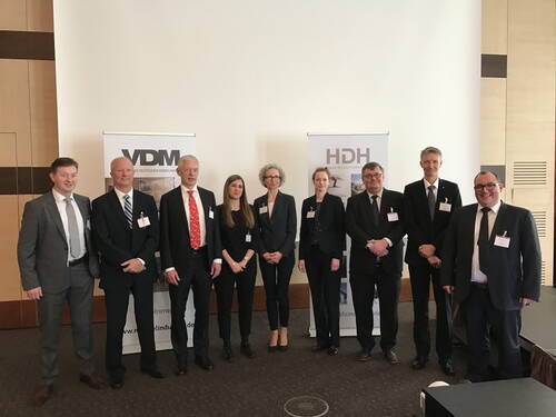 Neun Geschäftsleute in formeller Kleidung stehen nebeneinander vor Roll‑Up‑Bannern der Verbände VDM und HDH in einem Konferenzraum
