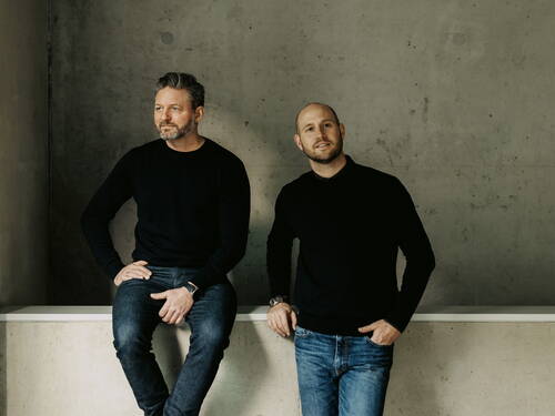 Zwei Männer in schwarzen Pullovern und Jeans posieren vor einer grauen Betonwand, einer sitzend und nach links blickend, der andere stehend und in die Kamera lächelnd.