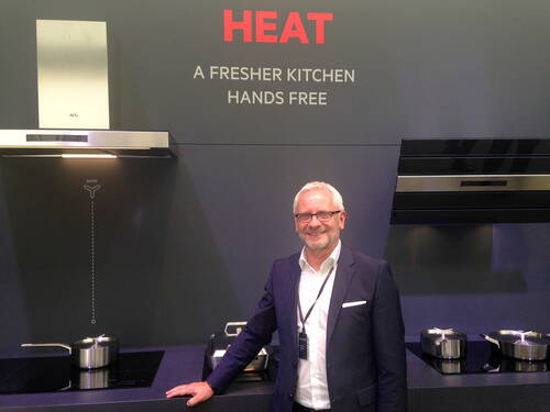 Lächelnder Mann im Anzug präsentiert moderne AEG-Dunstabzugshauben und Kochfelder vor einer dunkelgrauen Messewand mit dem Schriftzug „HEAT – A FRESHER KITCHEN HANDS FREE“.