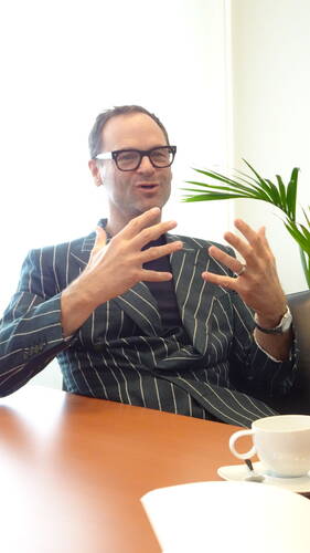 Mittelalter Mann im gestreiften Sakko sitzt an einem Konferenztisch, spricht lebhaft mit ausdrucksvollen Handbewegungen neben einer Kaffeetasse in einem hellen Büro.