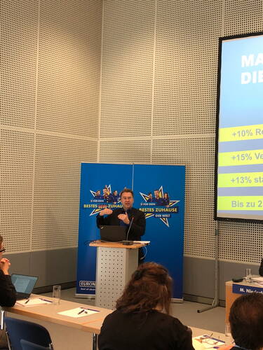 Referent präsentiert an einem Rednerpult vor blauem Werbehintergrund Marketingzahlen auf einer großen Leinwand in einem Konferenzraum