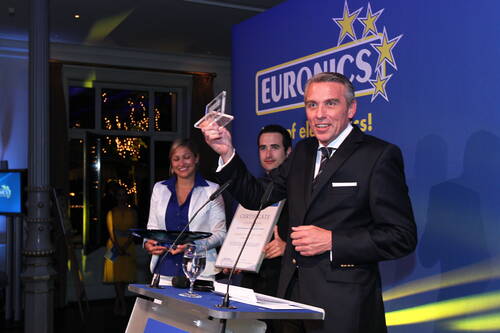 Geschäftsmann im Anzug hält strahlend einen transparenten Award in die Höhe, während zwei Kollegen auf einer Euronics-Bühne mit Urkunde und Tablet im Hintergrund stehen.
