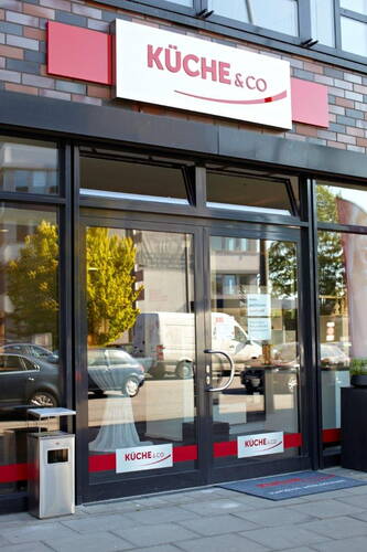 Eingang eines modernen Küche&Co-Fachgeschäfts mit großer Glasfront an einer Stadtstraße