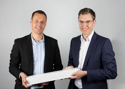 Zwei lächelnde Männer in Anzügen halten gemeinsam eine lange weiße Box vor neutralem Hintergrund