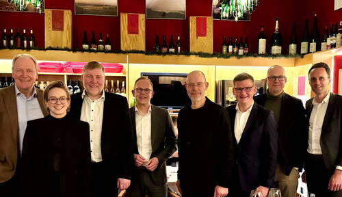Acht festlich gekleidete Personen stehen lächelnd nebeneinander in einer stimmungsvoll beleuchteten Weinbar vor Regalen mit Weinflaschen und Holzkisten.