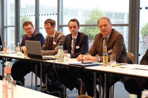 Mehrere Geschäftsleute im Anzug sitzen in einem Konferenzraum an einem langen Tisch, diskutieren vor einem Laptop und haben Getränke sowie Unterlagen vor sich.