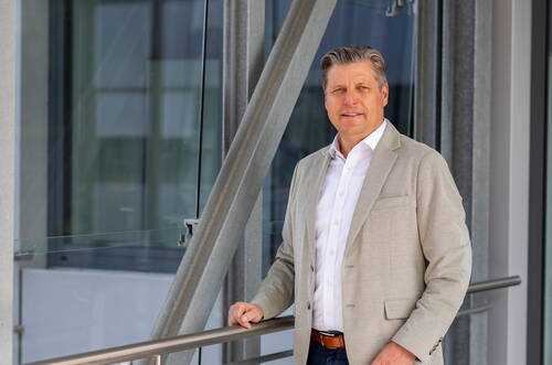 Geschäftsmann im beigen Sakko lehnt an einem Geländer in einem modernen Bürogebäude und blickt lächelnd in die Kamera