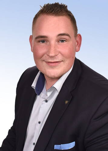 Junger Mann im dunklen Sakko und hellblauem Hemd lächelt freundlich vor hellem, verlaufendem Hintergrund