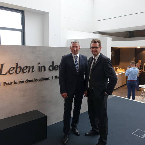 Zwei Männer im Anzug posieren lächelnd in einem modernen Ausstellungsraum vor einer Wand mit der Aufschrift „Leben in der Küche“, während im Hintergrund weitere Personen an einer Kücheninsel stehen.