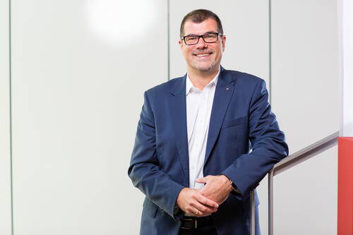 Lächelnder Mann im blauen Business-Anzug mit Brille steht an einem Treppengeländer in einem hellen, modernen Büro.