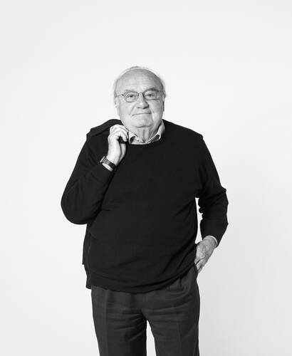 Älterer Mann mit Brille und dunklem Pullover steht vor hellem Hintergrund und hält lässig seinen Kragen mit einer Hand, während die andere in der Hosentasche ruht.