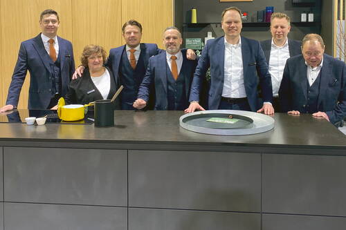Siebenköpfiges Team in Anzug und Businesskleidung steht lächelnd hinter einer modernen Kücheninsel mit gelbem Kochtopf und Dekotablett in einer Ausstellungsumgebung.