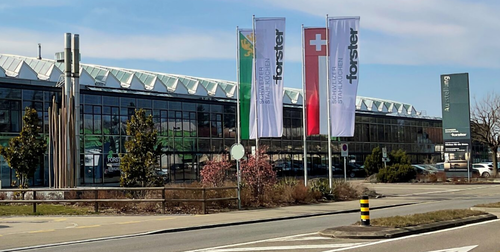 Moderne Glasfassade eines Forster-Ausstellungsgebäudes mit wehenden Schweizer und Firmenflaggen an einer mehrspurigen Straße bei sonnigem Wetter