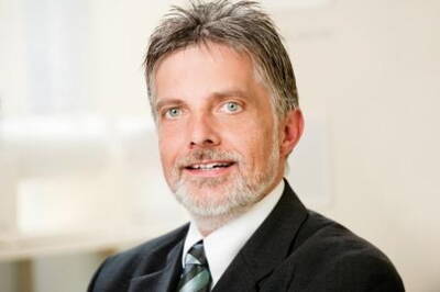Mittelalterlicher Mann mit graumelierten Haaren und Bart lächelt im Business-Anzug mit gestreifter Krawatte in einem hellen Büroraum in die Kamera.