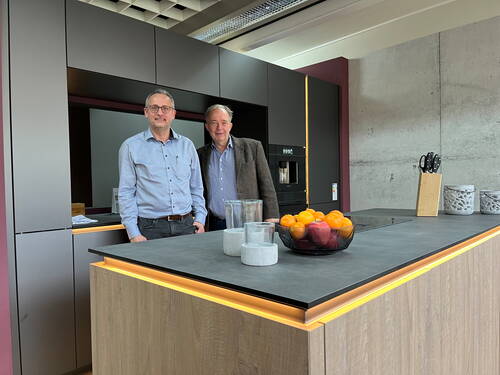 Zwei Männer stehen lächelnd hinter einer modernen Kücheninsel mit Schalen Obst, Marmorbehältern und Messerblock vor einer grauen Betonwand und dunklen Einbauschränken.