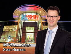 Junger Verkaufsleiter im Anzug steht lächelnd vor dem hell beleuchteten Eingangsbereich eines Küchenfachmarkts bei Nacht