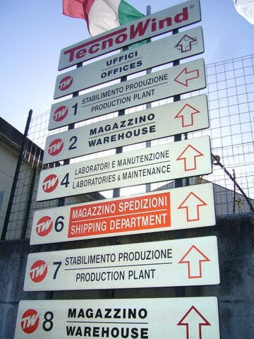 Wegweiser eines TecnoWind‑Industriegeländes mit nummerierten Hinweisschildern zu Büros, Produktionsstätten, Lagern, Laboren und Versandabteilung in Italienisch und Englisch