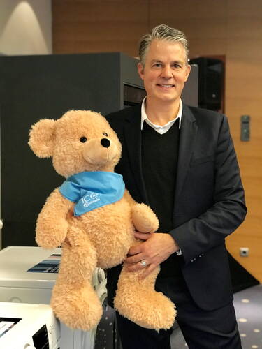Mittelalterlicher Mann im Anzug lächelt in moderner Büro- oder Ausstellungsumgebung und hält einen großen, hellbraunen Teddybären mit türkisblauem Halstuch im Arm