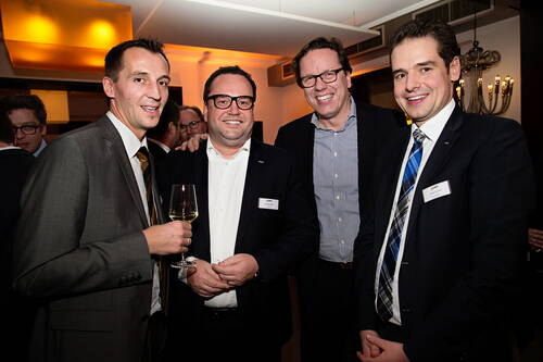 Vier lächelnde Geschäftsmänner in Anzügen stehen eng beisammen bei einer Abendveranstaltung, einer hält ein Weinglas, während im Hintergrund weitere Gäste in warm beleuchteten Räumen zu sehen sind.