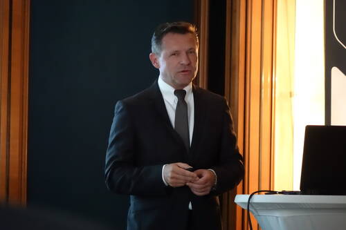 Mann im dunklen Anzug hält einen Vortrag und steht neben einem Pult mit Laptop in einem schwach beleuchteten Konferenzraum
