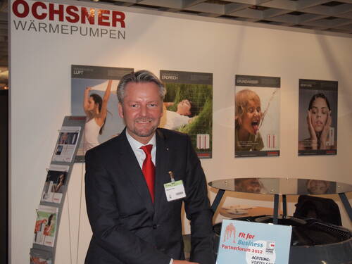 Geschäftsmann im Anzug präsentiert am Messestand von Ochsner Wärmepumpen ein Gerät neben einem „Fit for Business Partnerforum 2012“-Schild vor Werbeplakaten an der Wand