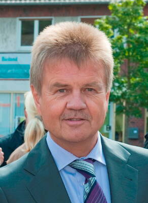 Mittelalterlicher Mann im Anzug mit Krawatte blickt direkt in die Kamera, während im Hintergrund Menschen und Gebäude einer Innenstadt zu sehen sind.