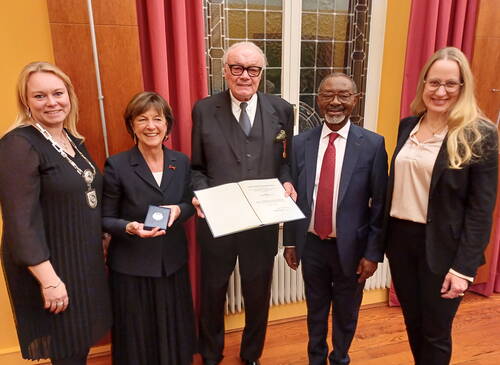 Fünf festlich gekleidete Personen stehen in einem repräsentativen Saal, während die Frau in der Mitte eine Medaille und der ältere Herr neben ihr eine geöffnete Ehrenurkunde präsentieren.