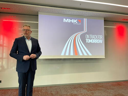 Business-Redner im Anzug präsentiert vor einer Leinwand mit dem MHK-Group-Logo und dem Slogan „On Track for Tomorrow“ in einem modern beleuchteten Konferenzraum