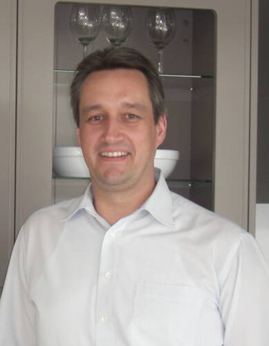 Mittelalterlicher Mann mit hellem Hemd lächelt in einer modernen Küche vor einem Schrank mit Gläsern und Schalen