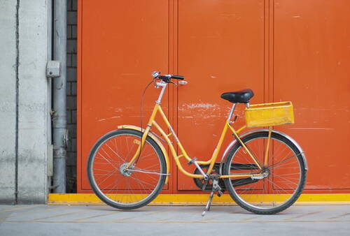 Gelbes Cityrad mit Gepäckkorb lehnt vor einem großen orangefarbenen Garagentor in einer Industrieumgebung.