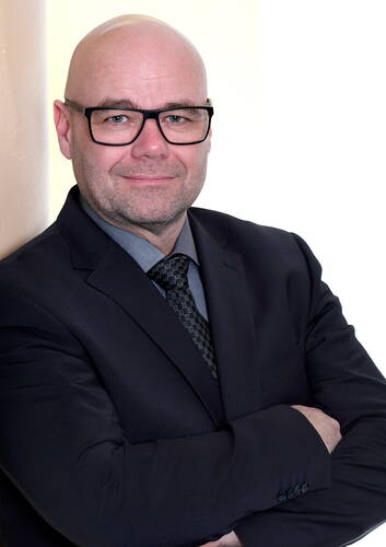 Glatzköpfiger Mann mit Brille im dunklen Anzug und gemusterter Krawatte steht mit verschränkten Armen vor hellem Hintergrund und blickt ruhig in die Kamera