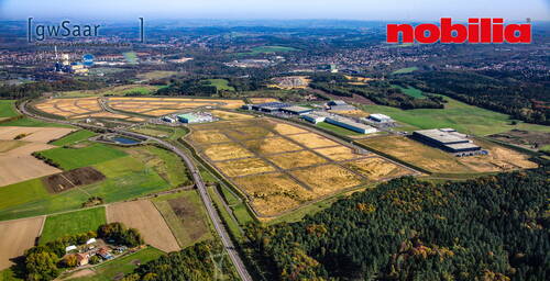Luftaufnahme eines weitläufigen Industrie- und Gewerbegebiets mit mehreren Hallen, Erschließungsstraßen und umgebenden Feldern am Stadtrand in hügeliger Landschaft