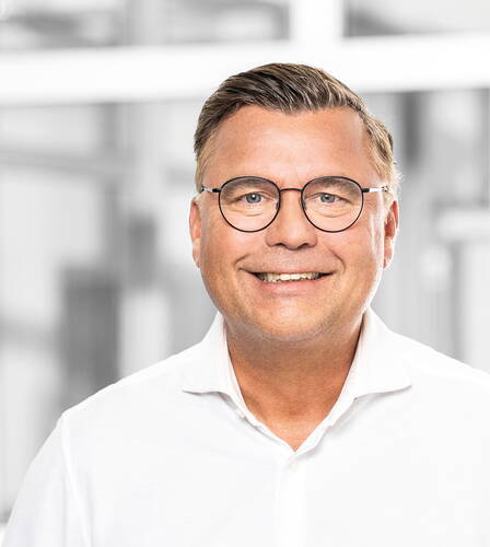 Lächelnder Mann mit Brille in weißem Hemd steht vor einem hellen, unscharfen Bürohintergrund