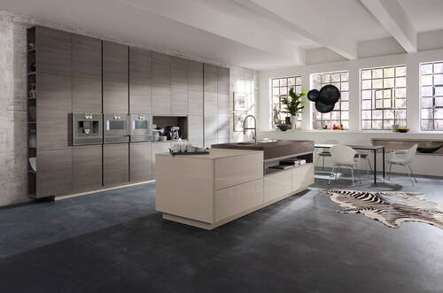Großzügige, lichtdurchflutete Designküche mit Kochinsel, wandhohen Holzfronten, integrierten Edelstahlgeräten, Essbereich am Fenster und auffälligem Zebrafell-Teppich auf dunklem Boden