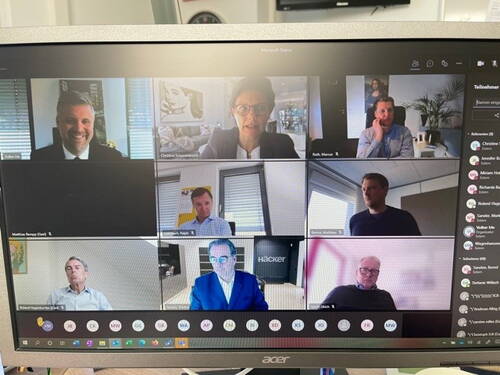Mehrere Geschäftsleute nehmen über Microsoft Teams an einer Videokonferenz teil, auf einem Monitor mit geöffneter Teilnehmerliste und verschiedenen Büroumgebungen im Hintergrund.
