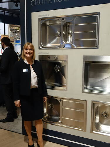 Geschäftsfrau im Anzug präsentiert an einem Messestand eine Wand mit mehreren glänzenden Edelstahl-Küchenspülen des Herstellers Grohe.